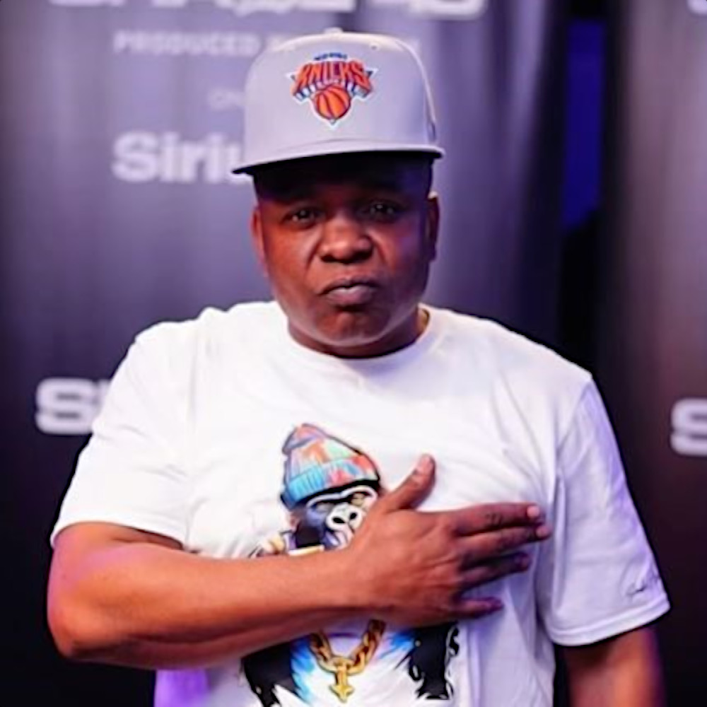 American rapper Snyp Life of D-Block standing snarling at the camera with his right hand on his chest in the shape of a pistol wearing a New York Knicks baseball cap and a T-shirt with a cartoon gorilla, colour image.
