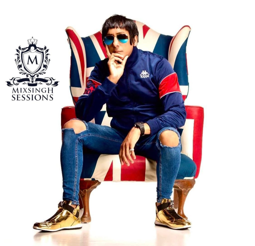 British DJ and producer Chelsea Singh sitting in a United Kingdom flag chair wearing sunglasses and a blue Kappa tracksuit top and wearing jeans with his Mixsingh Sessions logo next to him, colour image.