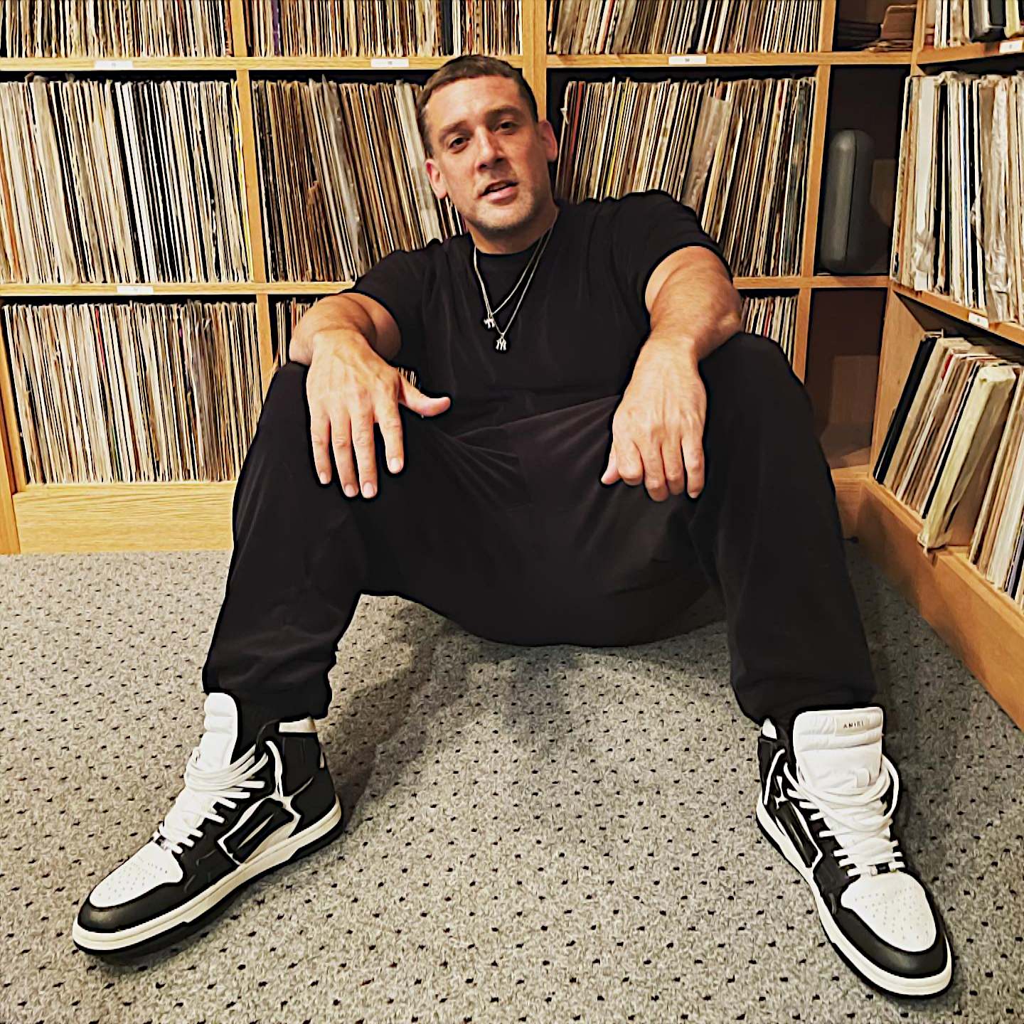 American DJ, emcee, and producer Scram Jones sitting on the floor in his vinyl vault with his arms on his knees leaning back on the vinyl records and smiling, colour image.