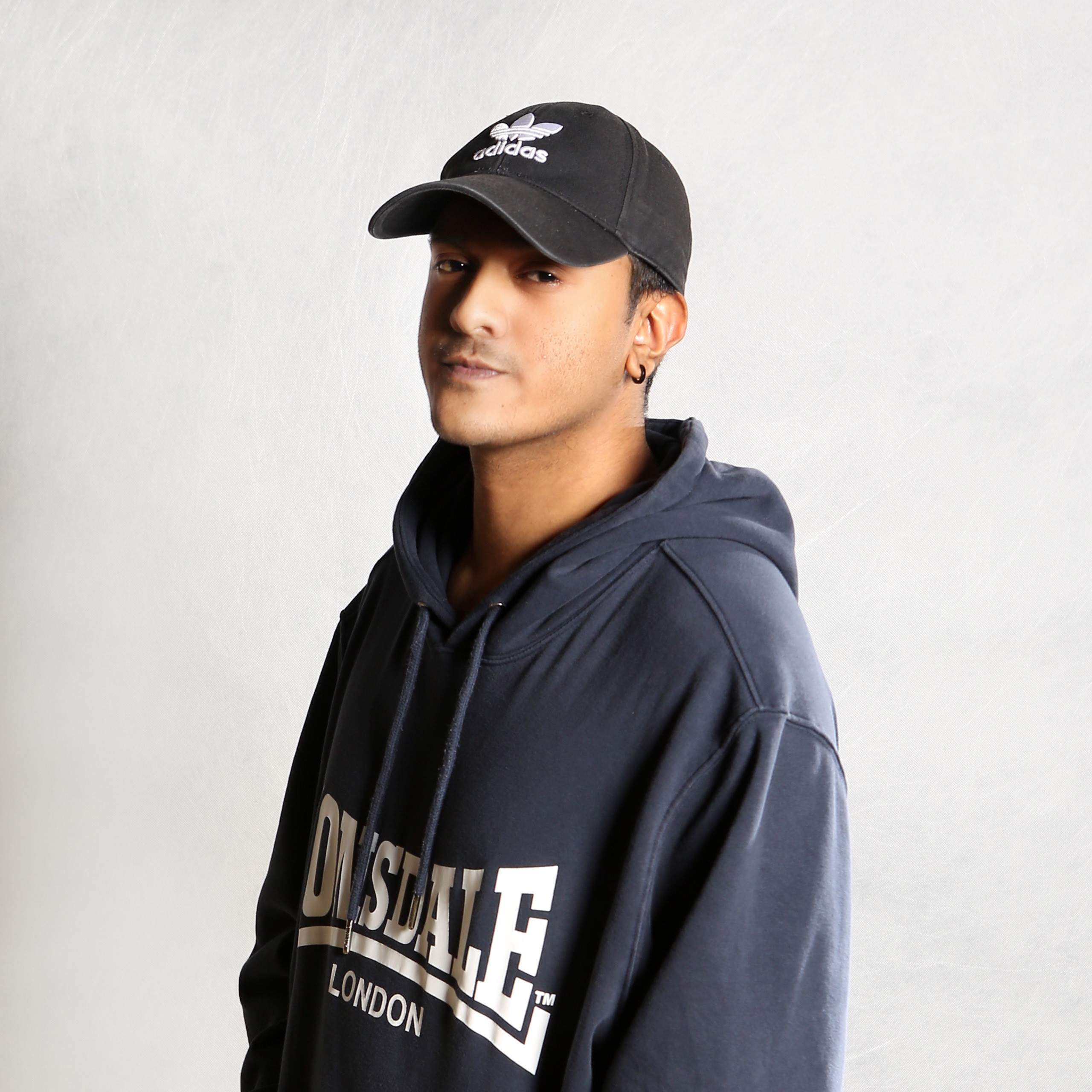 Malaysian DJ and producer St4n standing in a photographic studio wearing a Lonsdale hoodie and an Adidas baseball cap looking over his right shoulder at the camera, colour image.