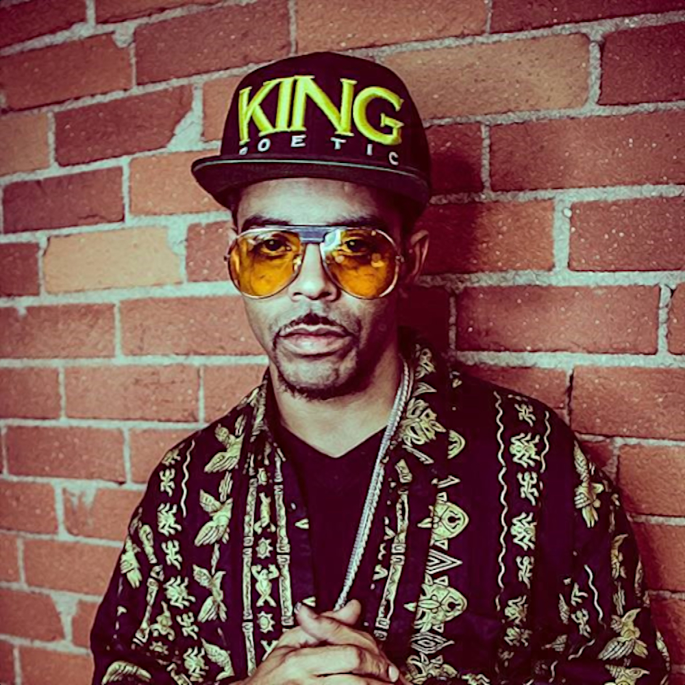 American rapper and producer Agallah standing against a brick wall wearing a King Poetic baseball cap and sunglasses looking at the camera with his hands together, colour image.