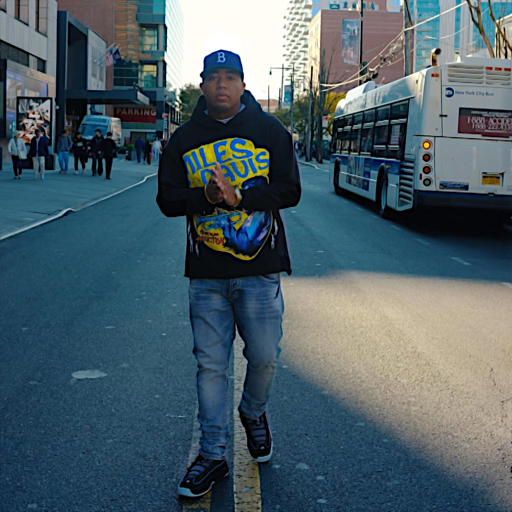 American rapper Skyzoo walking down the middle of a street in New York wearing a hoodie and baseball cap with his hands together and a New York City Bus behind his left side, colour image.