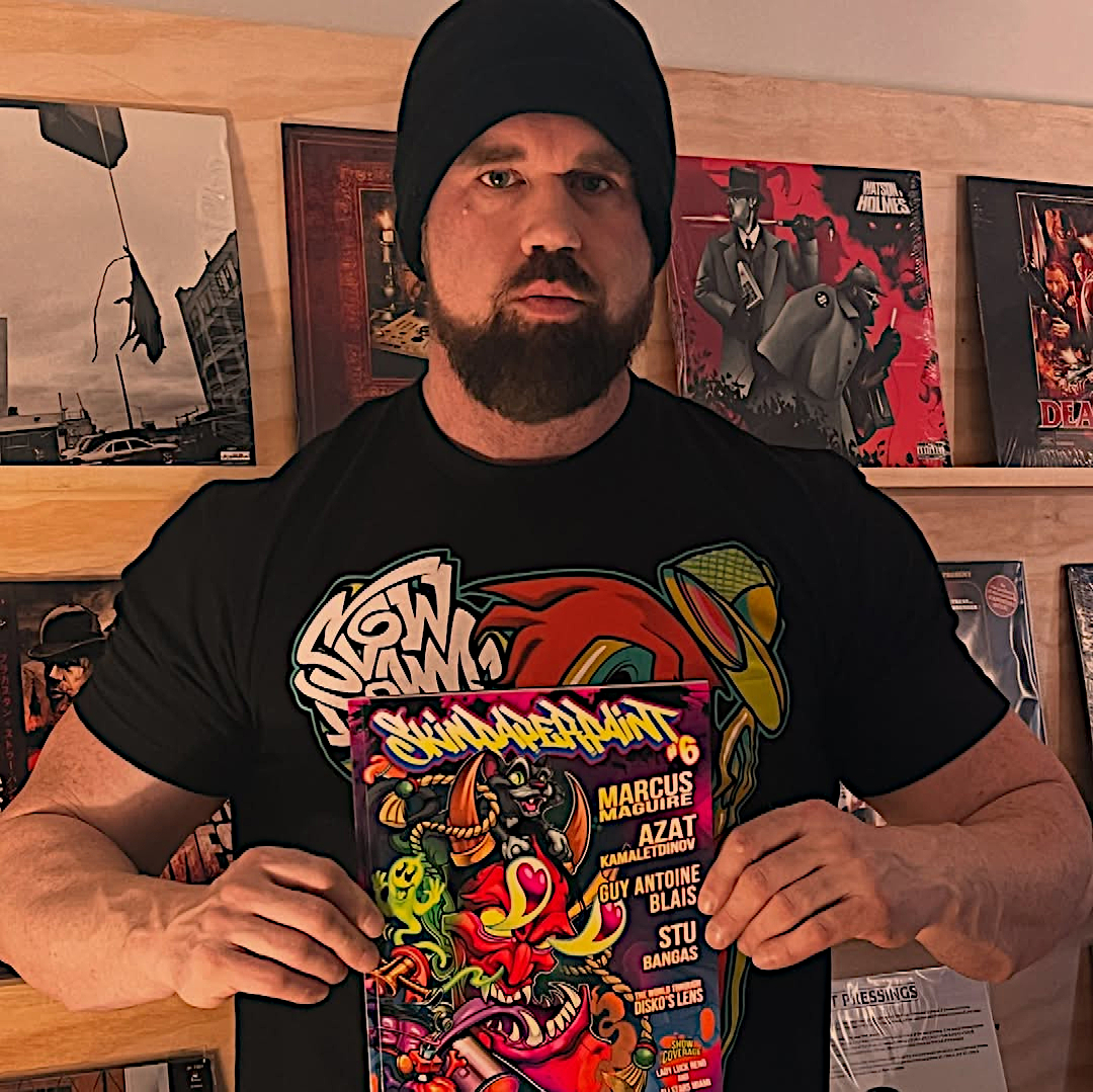 American hip hop producer Stu Bangas holding a magazine in front of himself, standing in a room with art behind him wearing a beenie hat, colour image.
