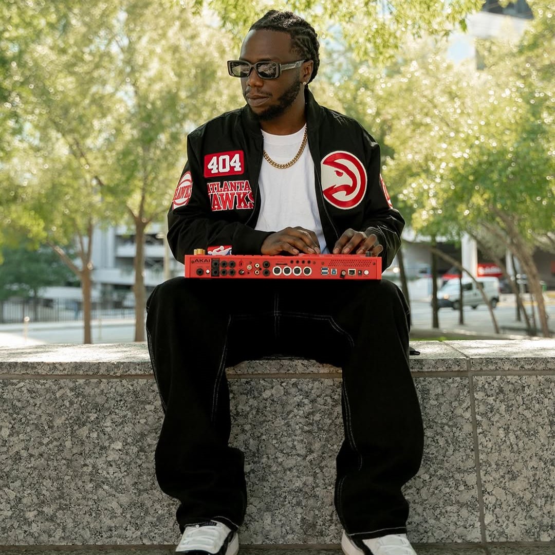 American hip-hop producer BeatsByJBlack sitting wearing sunglasses and an Atlanta Hawks Jacket in a park on a wall with an Akai drum machine on his lap, colour image.