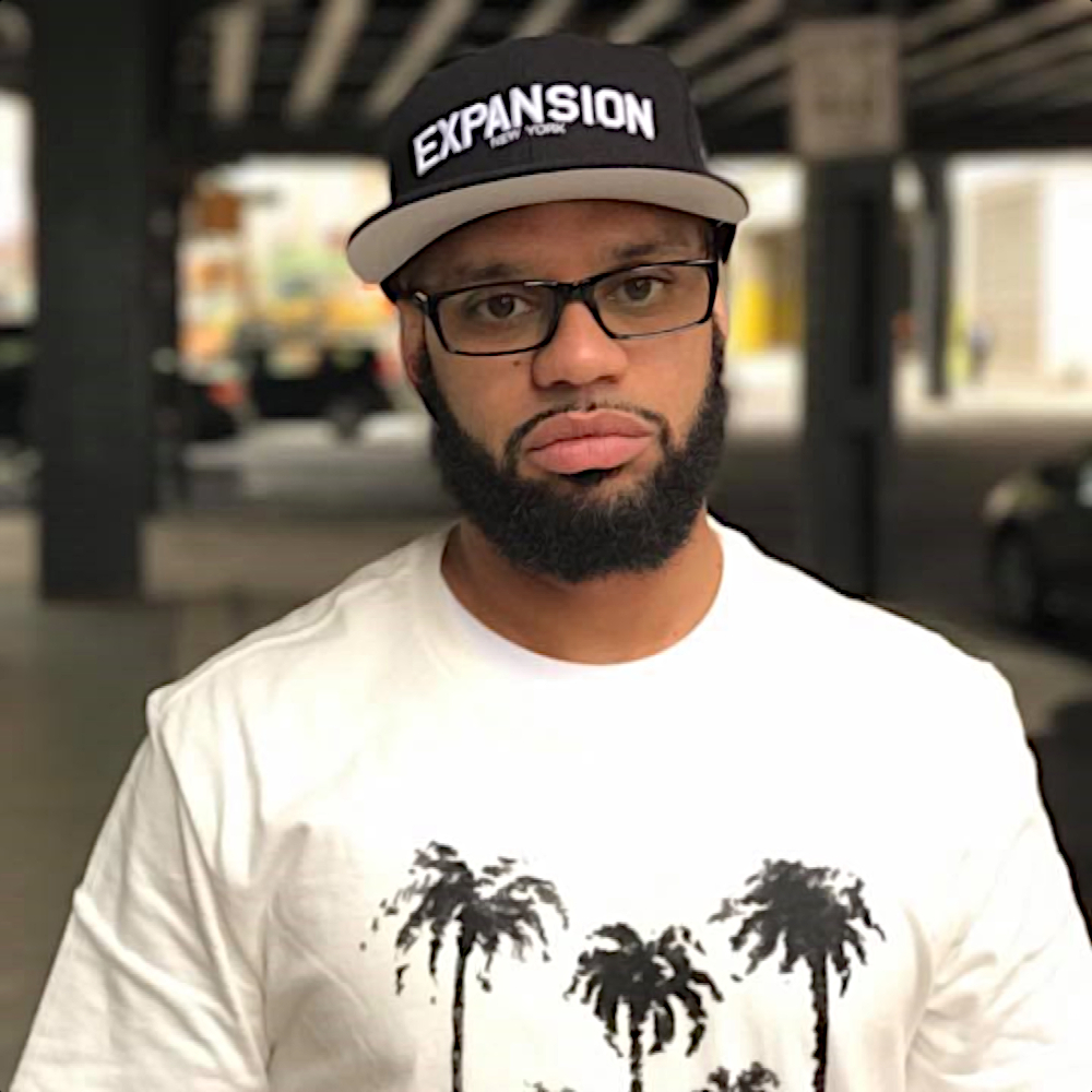 American hip-hop rapper Nutso at a video shoot wearing a baseball cap with expansion written on it and a T-shirt with some palm trees, standing under a subway station, colour image.