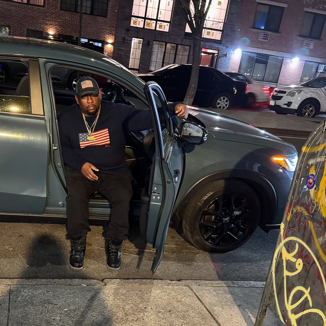American rapper Shabaam Sahdeeq sitting in the door of his car with the door open on a street in Brooklyn, New York, USA wearing a Ralph Lauren sweater and baseball cap, colour image