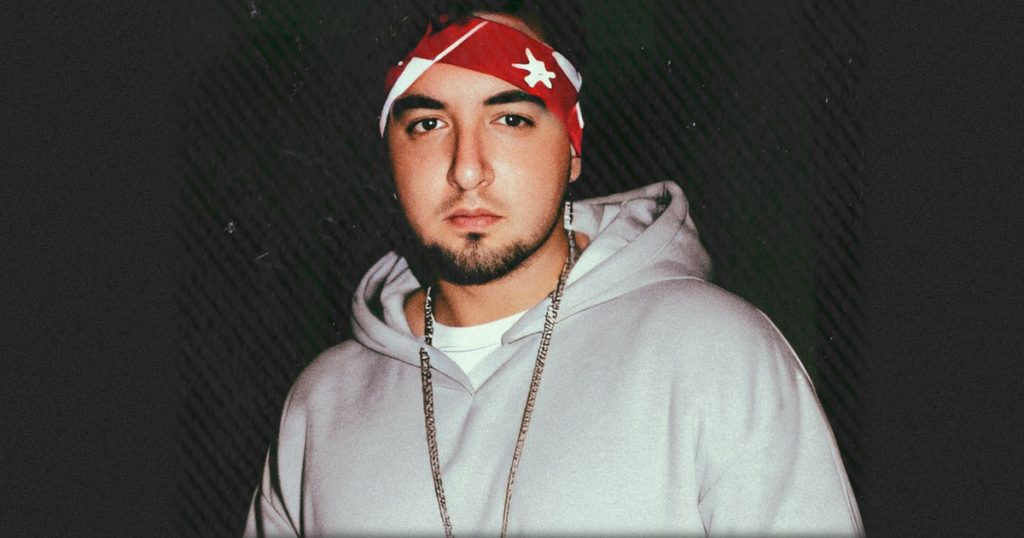 Italian DJ and producer Daniel Kaiser Malengo standing wearing a grey hoodie and red bandana on his head looking at the camera, colour image with black background.