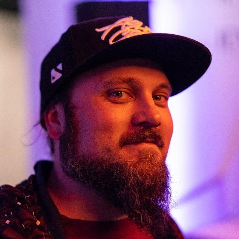 French Canadian Rapper and Artist Monk.E wearing a baseball cap and smirking whilst facing left at 45 degrees and looking at the camera, colour image.