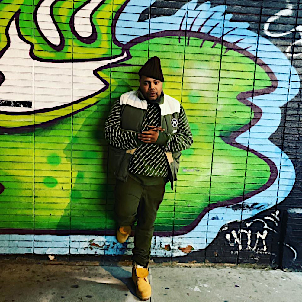 American rapper and producer John Jigg$ (John Jiggs) leaning against a graffiti wall in the street with his right foot on the wall and his hands together in front of him wearing a padded vest, sweater and jeans with Timberland boots, colour image.