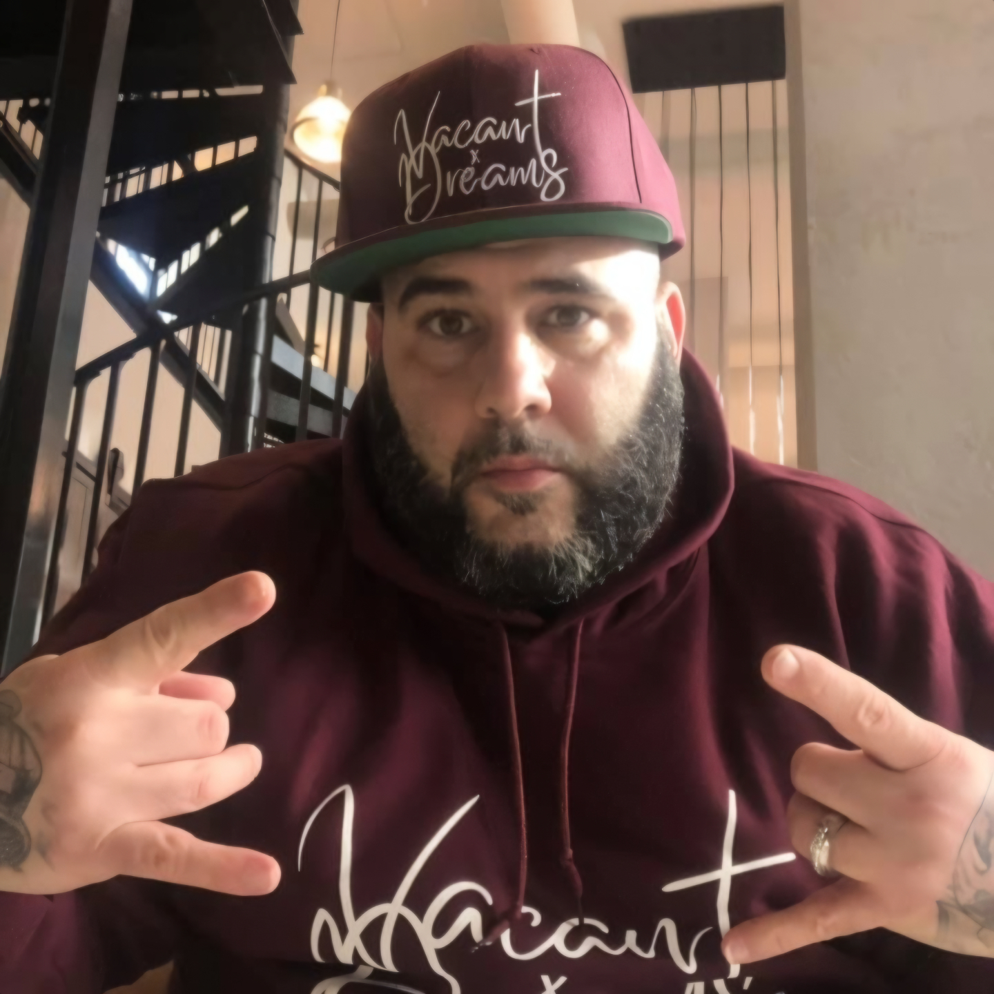 American hip-hop rapper Jus One wearing a Vacant Dreams hoodie and Baseball Cap with his fingers in an inward devil's horn gesture with some spiral stairs behind him, colour image