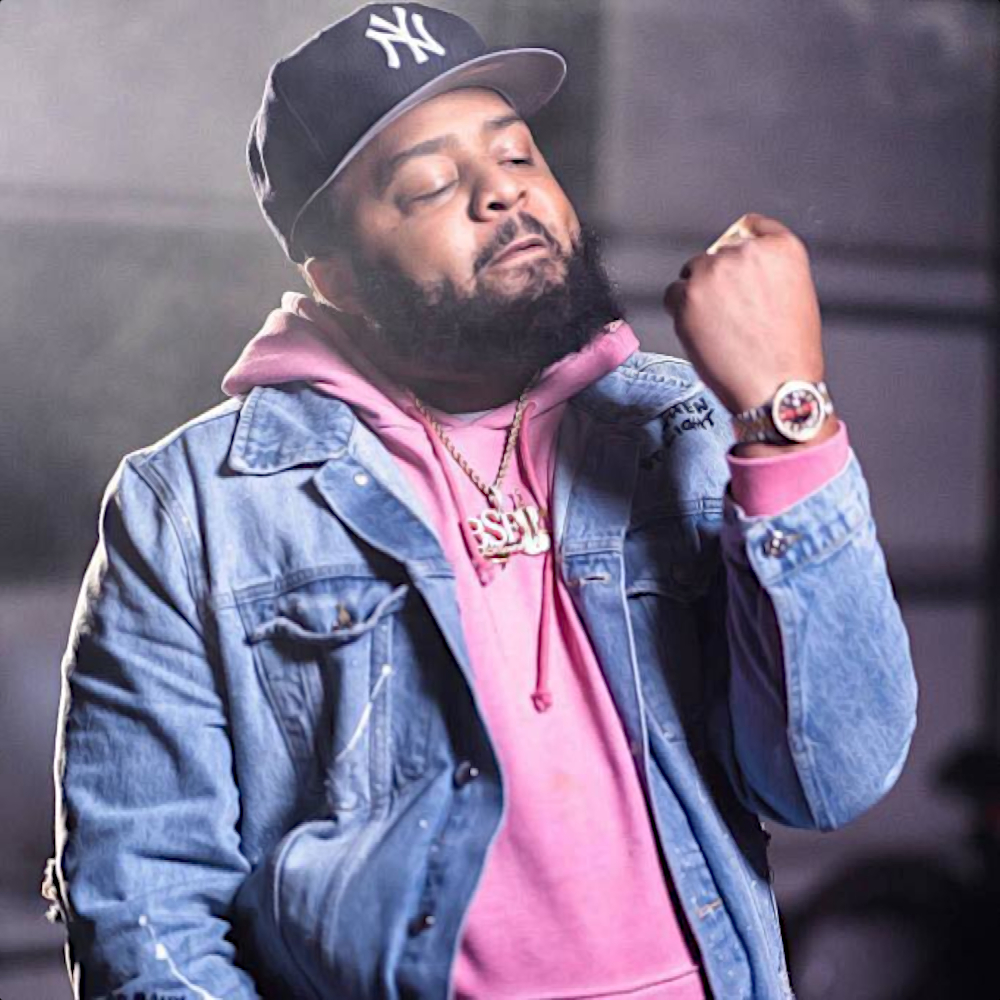 American hip-hop rapper and producer Rick Hyde, looking at his left fist wearing a Yankees baseball cap and a denim jacket over a pink hoodie, colour image.