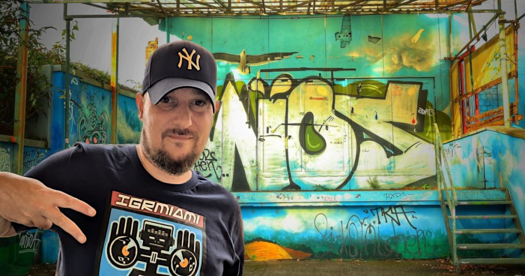 French DJ Phoenixx standing in front of a derelict stage with wall covered in Graffiti in Germany wearing a Yankees baseball cap and holding his fingers in a two fingered salute, colour image.