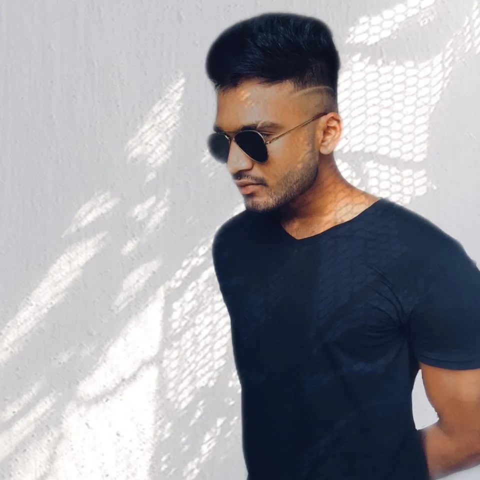 Hersiaz Hazi wearing sunglasses and a black t-shirt stands against a textured wall with shadows cast by a net-like pattern.