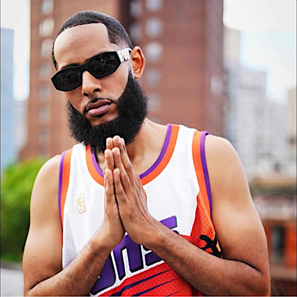 Ca sĩ rap hip-hop người Mỹ The Musalini mặc áo bóng chày và đeo kính râm, hai tay chắp lại cầu nguyện trên đường phố ở New York, phía sau là một khu chung cư; hình ảnh màu.
