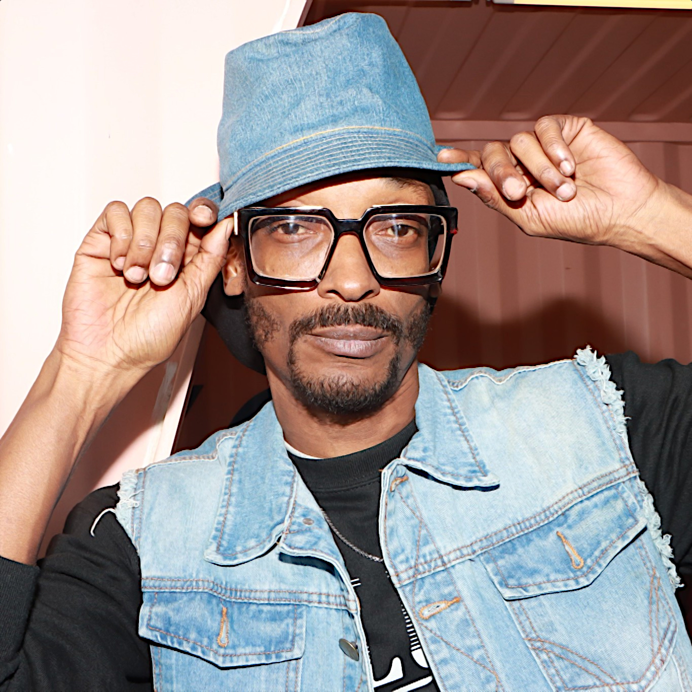 Rapper và nhà sản xuất người Mỹ 4-IZE đứng, mặc áo ghi-lê denim, đội mũ và đeo kính, hai tay đặt lên vành mũ, đang nhìn vào máy ảnh, hình màu.