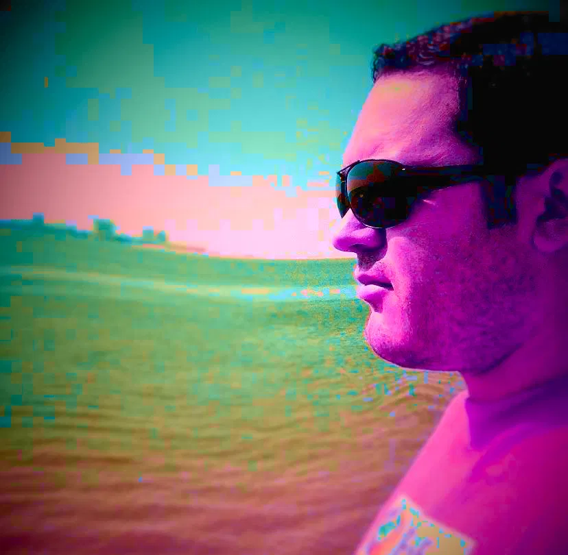Portuguese DJ Joshua standing 0on a beach facing left with the sea in the background, pink effect, colour image.