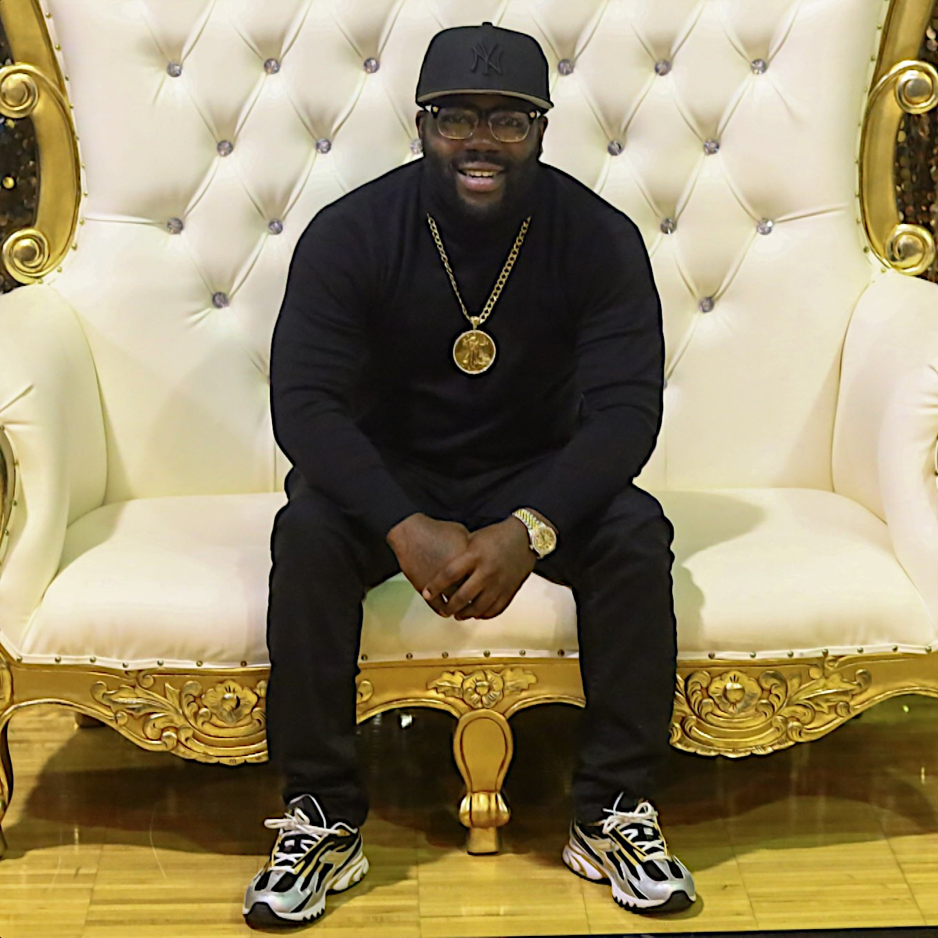 American rapper Jamal Gasol sitting on a luxury chair wearing a baseball cap, glasses, a gold chain, black jeans, and a black sweater in a colour image