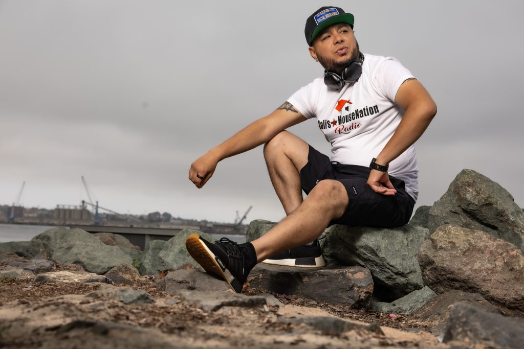 American DJ and radio presenter DJ Nocturne sitting on some rocks close to the sea looking over his left shoulder and wearing a baseball cap, T-shirt, and shorts with his right arm on his right knee, colour image