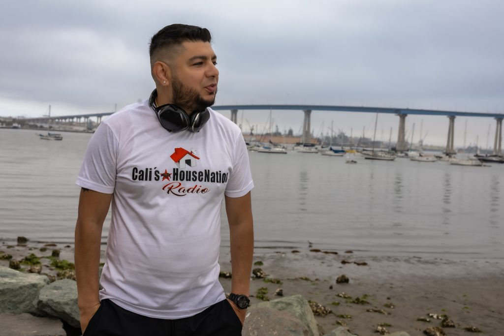 American DJ and radio presenter DJ Nocturne standing close to the sea looking over his left shoulder and wearing a baseball cap, T-shirt, and shorts with his right arm on his right knee, colour image.