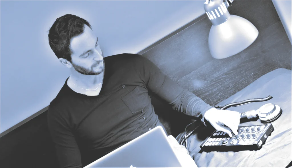 Italian DJ and producer Inve sitting on a bed with his laptop on his lap playing with a music sampler, black and white image with blue tinge.