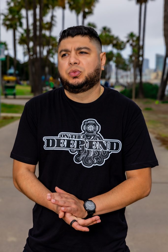 American DJ and radio presenter DJ Nocturne standing in a park with his hands held together at the palms in front of him and wearing a black On The Deep End T-shirt, colour image.
