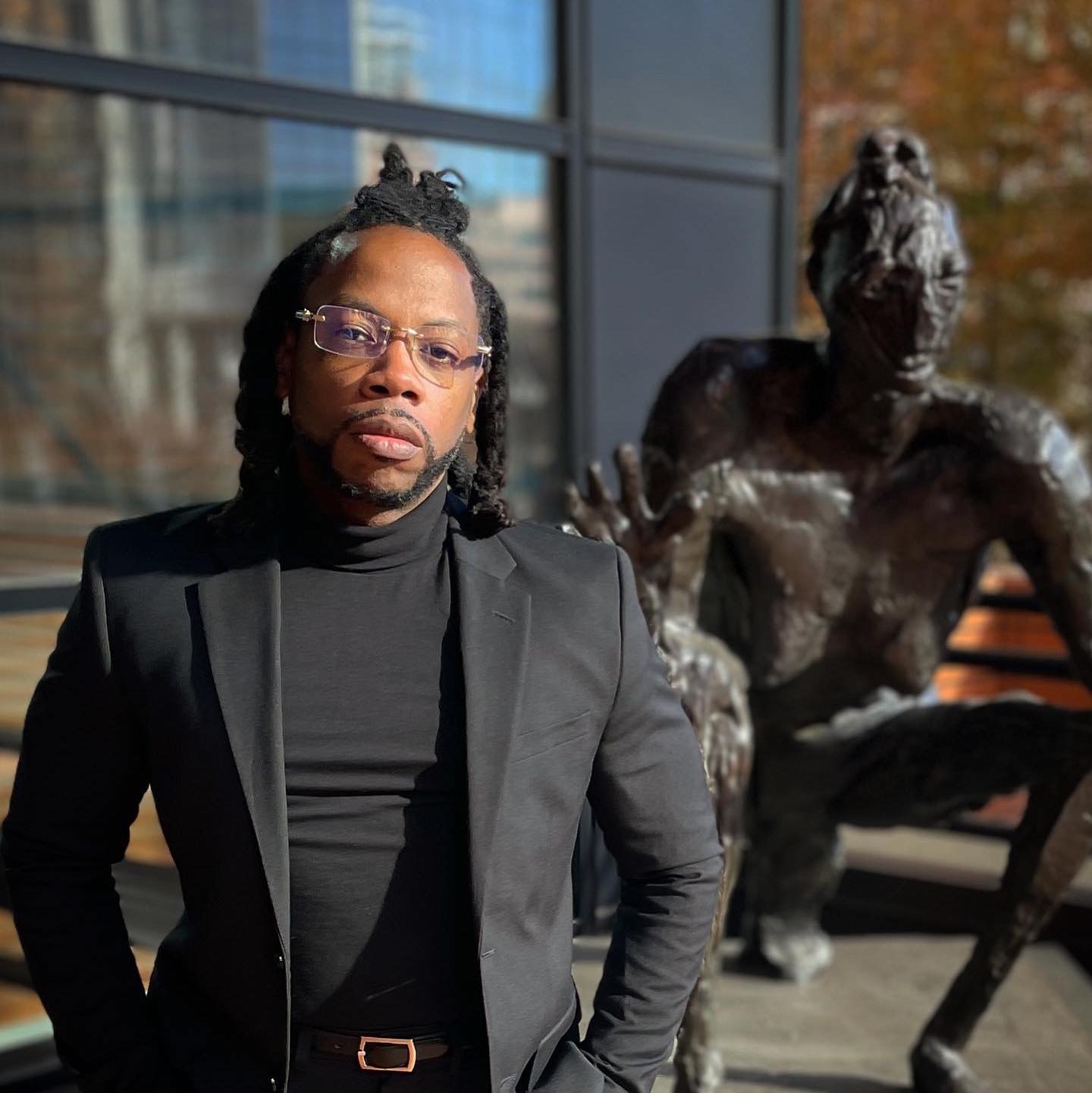 American rapper J-Hood standing in a suit with a roll-neck shirt underneath, glasses, and a short trimmed beard, his hair styled in short dreadlocks, posing in front of a statue in a color image.