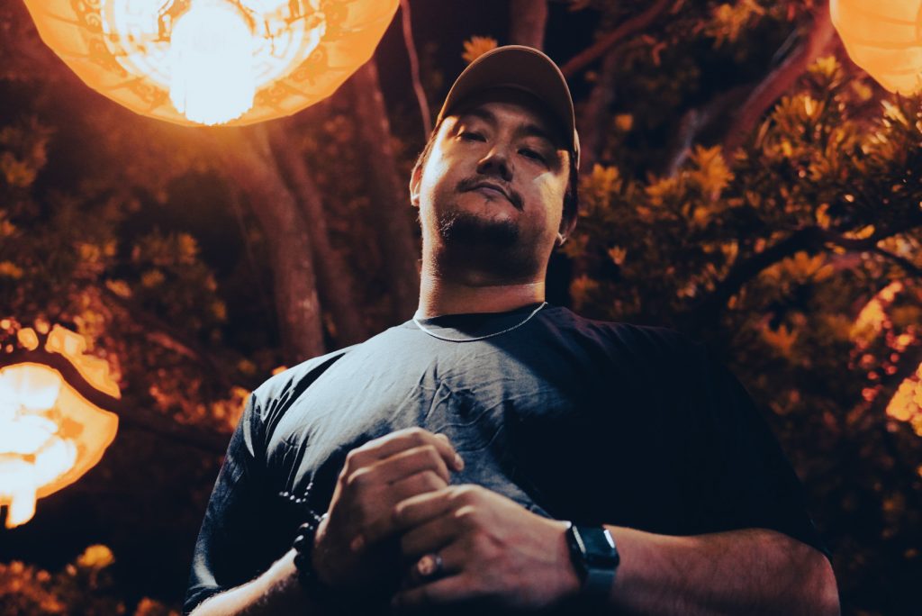 DZRT FRST, a Taipei DJ, stands next to a tree with a lantern, embodying his exploration of minimal house and house music in a colour image.