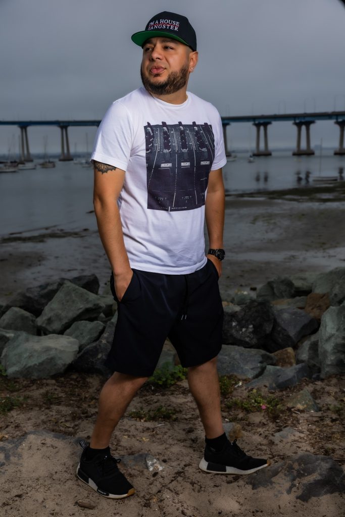 American DJ and radio presenter DJ Nocturne standing on a beach with his hands in the pockets of his shorts and wearing a baseball cap, sneakers, T-shirt and shorts, looking over his right shoulder in a colour image.