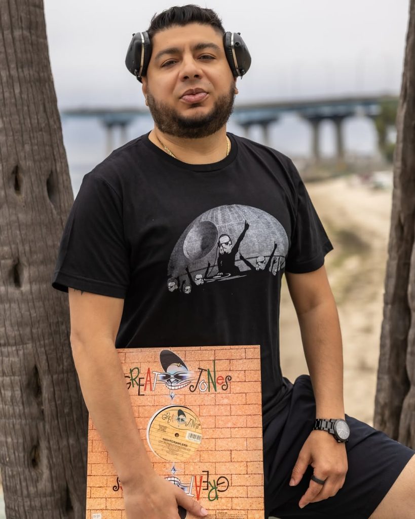 American DJ and radio presenter DJ Nocturne stands in a park, holding a vinyl record in his right hand, positioned between two trees, and wearing a black Star Wars Death Star T-shirt. The image is in color.