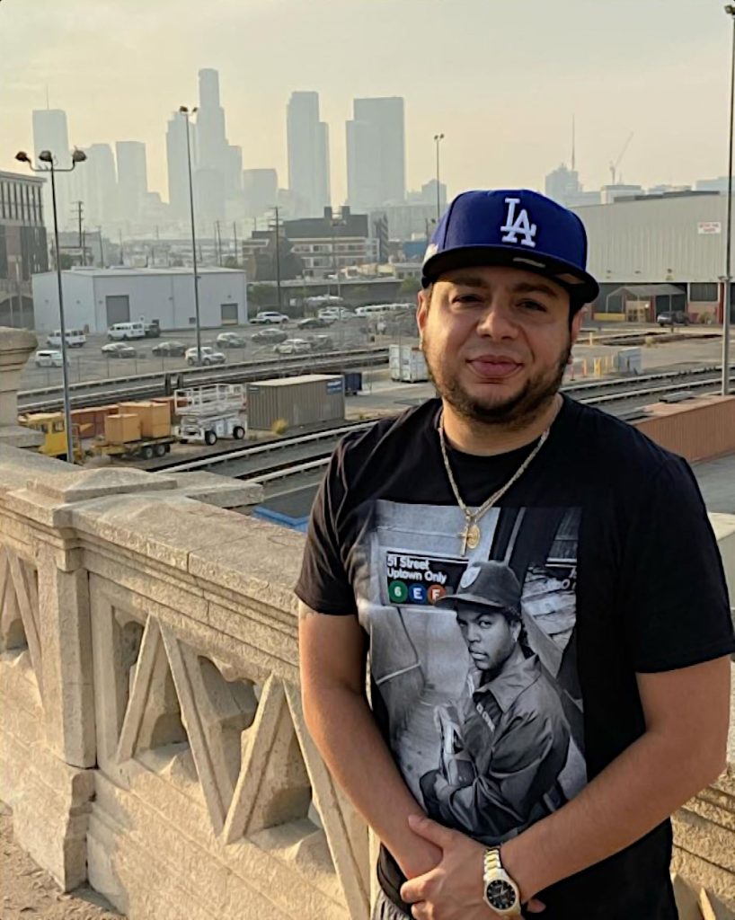 American DJ and radio presenter DJ Nocturne stands on a bridge walkway with an industrial estate in the background in, Los Angeles, California, USA, and wearing a black Easy-E of N.W.A T-shirt. The image is in color.