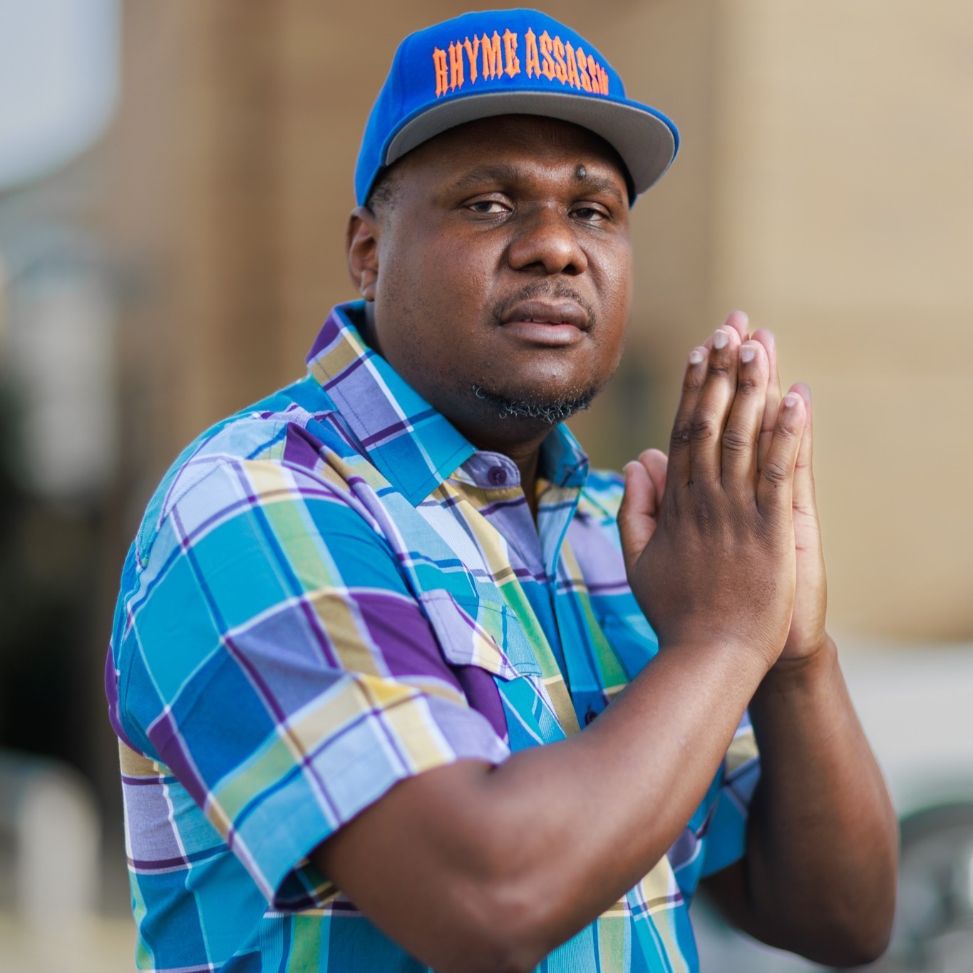 UK-based Zimbabwean rapper and record label owner Rhyme Assassin is positioned facing to the right, wearing a tartan short-sleeved shirt and a Rhyme Assassin baseball cap. He has his hands together in front of him in a prayer gesture while looking at the camera in a colourful image.