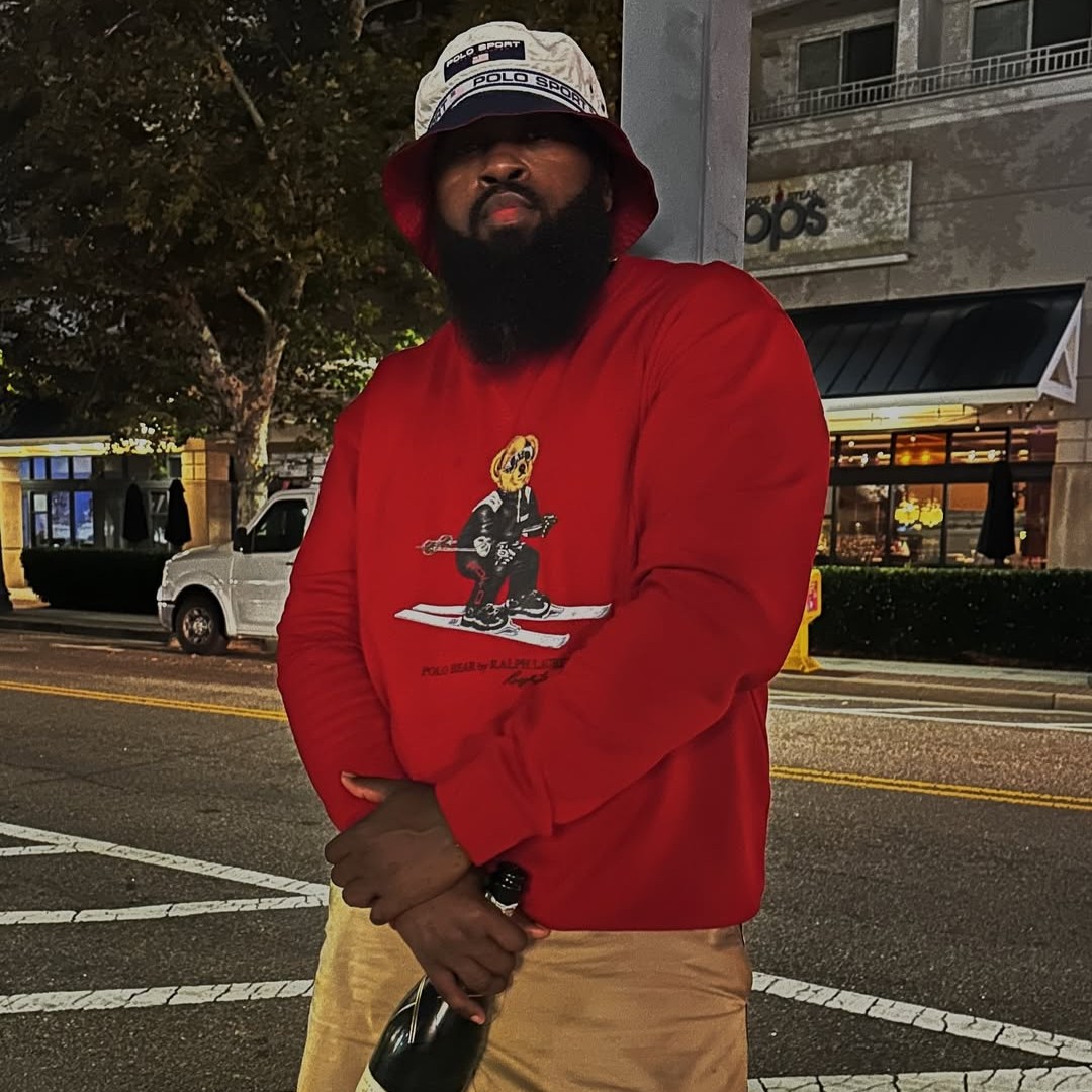 American hip-hop producer and rapper JR Swiftz holding a bottle of champagne in his right hand, wearing a red Polo Bear sweater and a Kangol-style hat, looking at the camera while standing in a street in a color image.