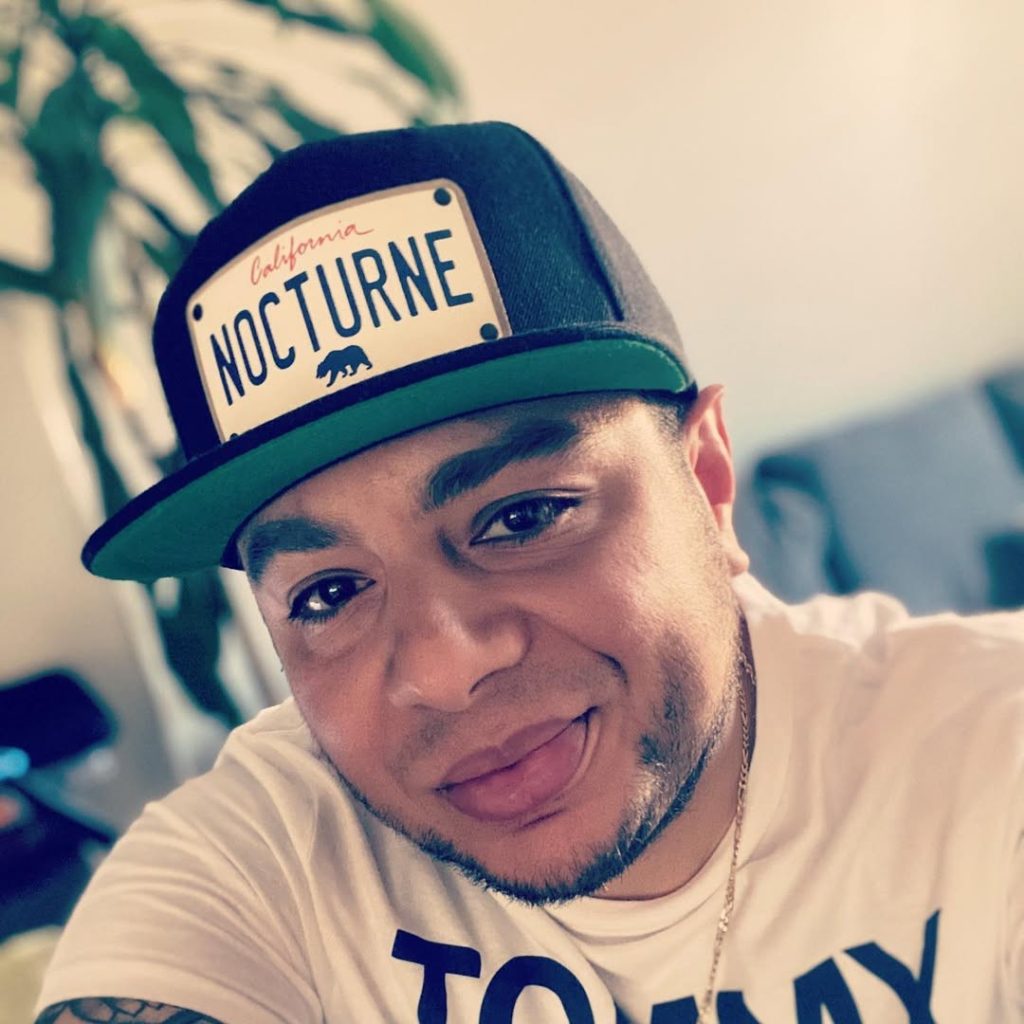 American DJ and radio presenter DJ Nocturne is captured in a color facial portrait, looking at the camera with a wry smile while wearing a Nocturne baseball cap. The photo is set in Los Angeles, California, USA.