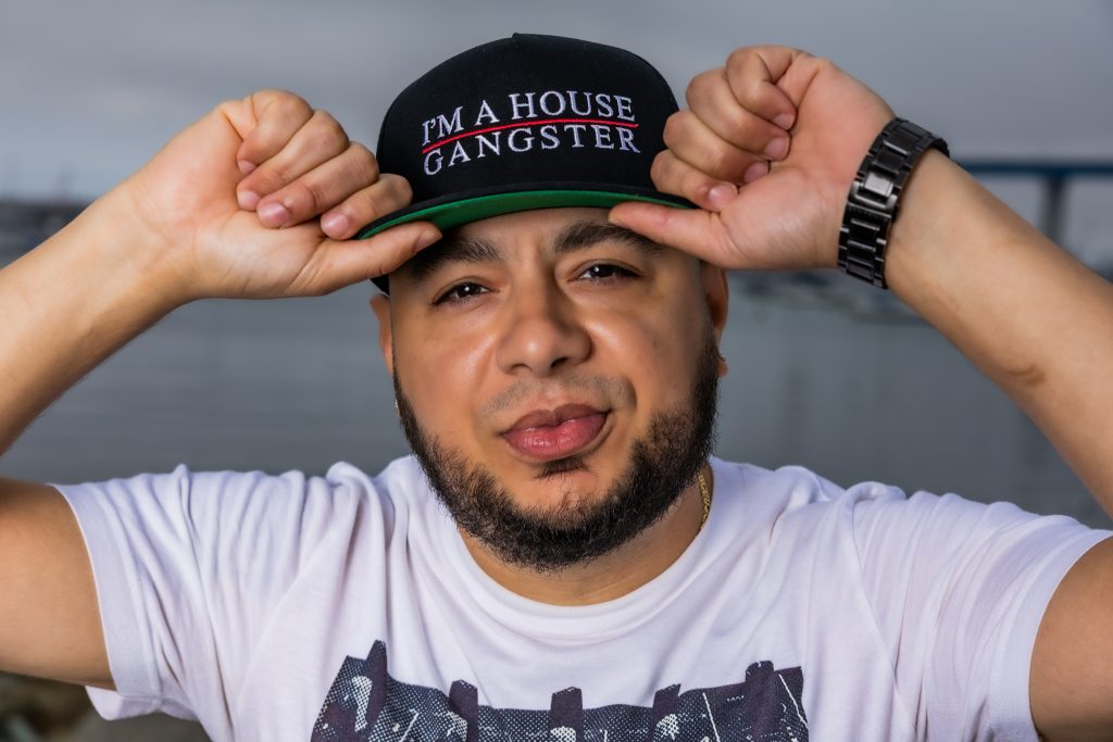 American DJ and radio presenter DJ Nocturne is featured in a colorful portrait, gazing at the camera with a wry smile. He wears an "I'm a House Gangster" baseball cap, holding the brim with both hands. The photo is set in Los Angeles, California, USA