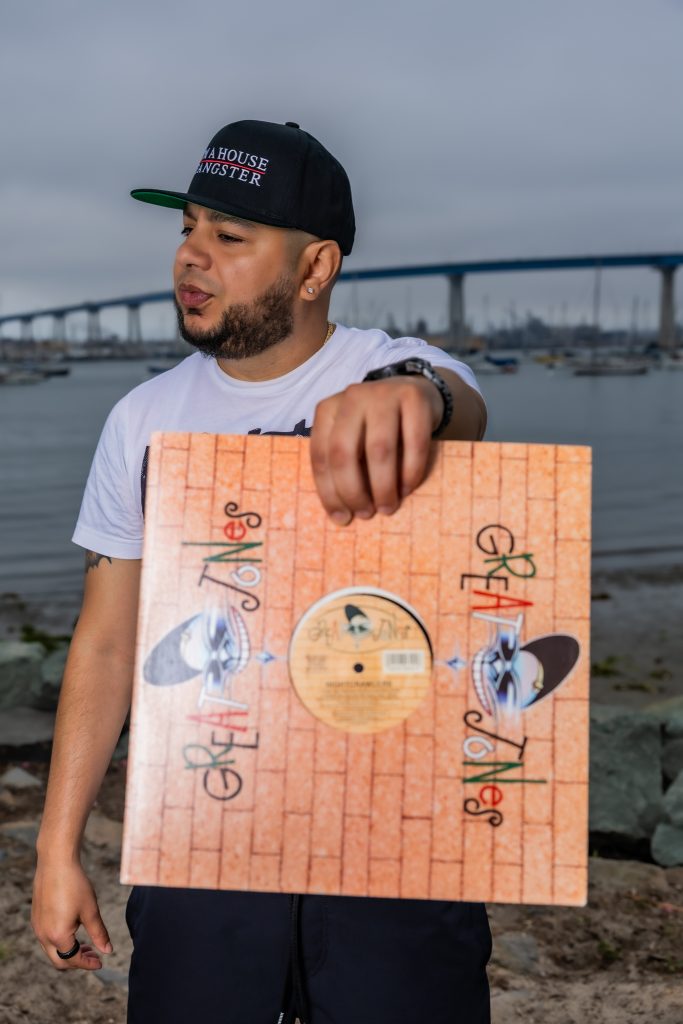 American DJ and radio presenter DJ Nocturne is captured in a vibrant portrait, gazing to his right while holding a vinyl record in his left hand. He sports an "I'm a House Gangster" baseball cap, with the scene set in Los Angeles, California, USA.