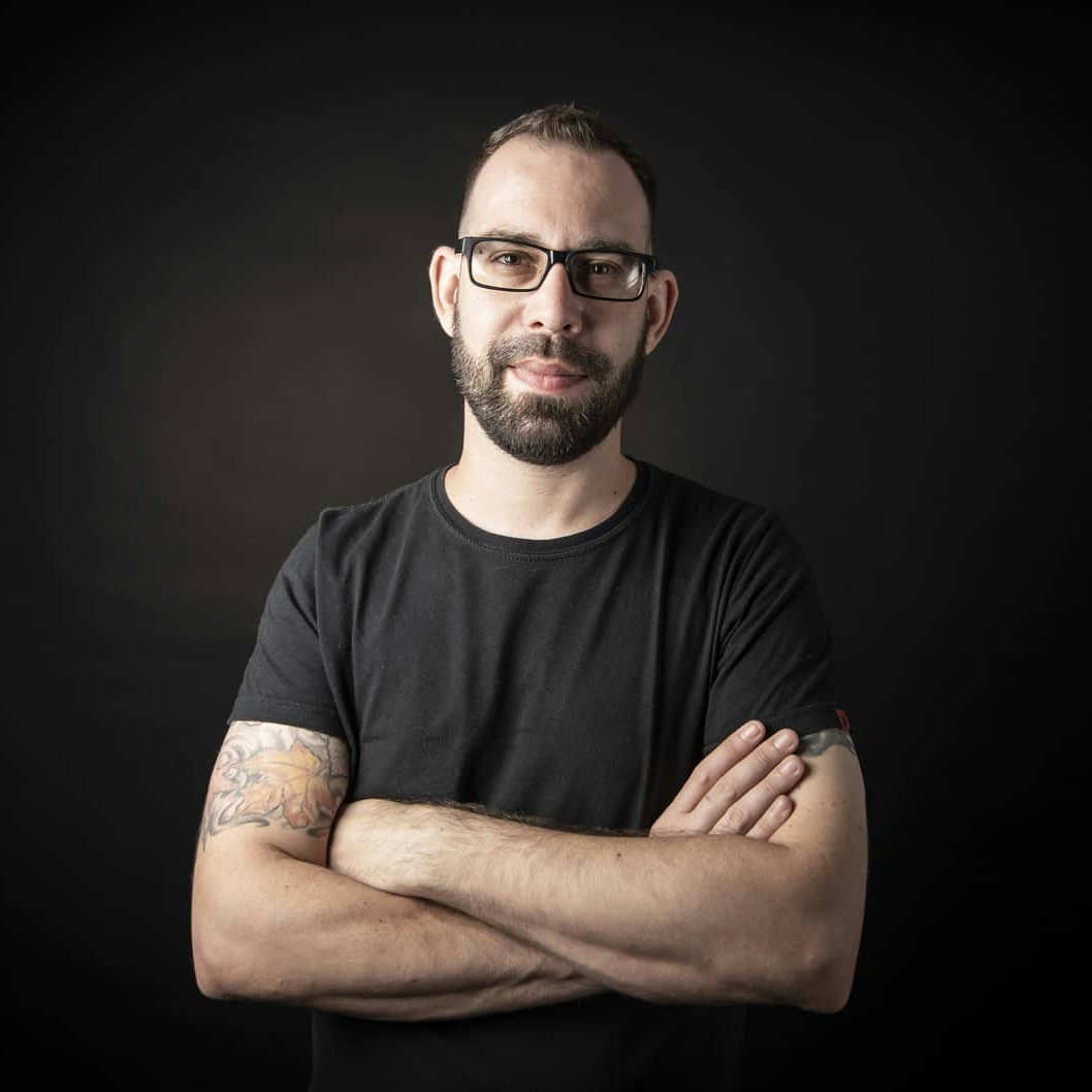Argentinian DJ and producer Kiky Pop stands in a studio, smirking at the camera with his arms crossed. He wears a black t-shirt, glasses, and a neatly trimmed short beard, captured in a color shot against a black and gray background.