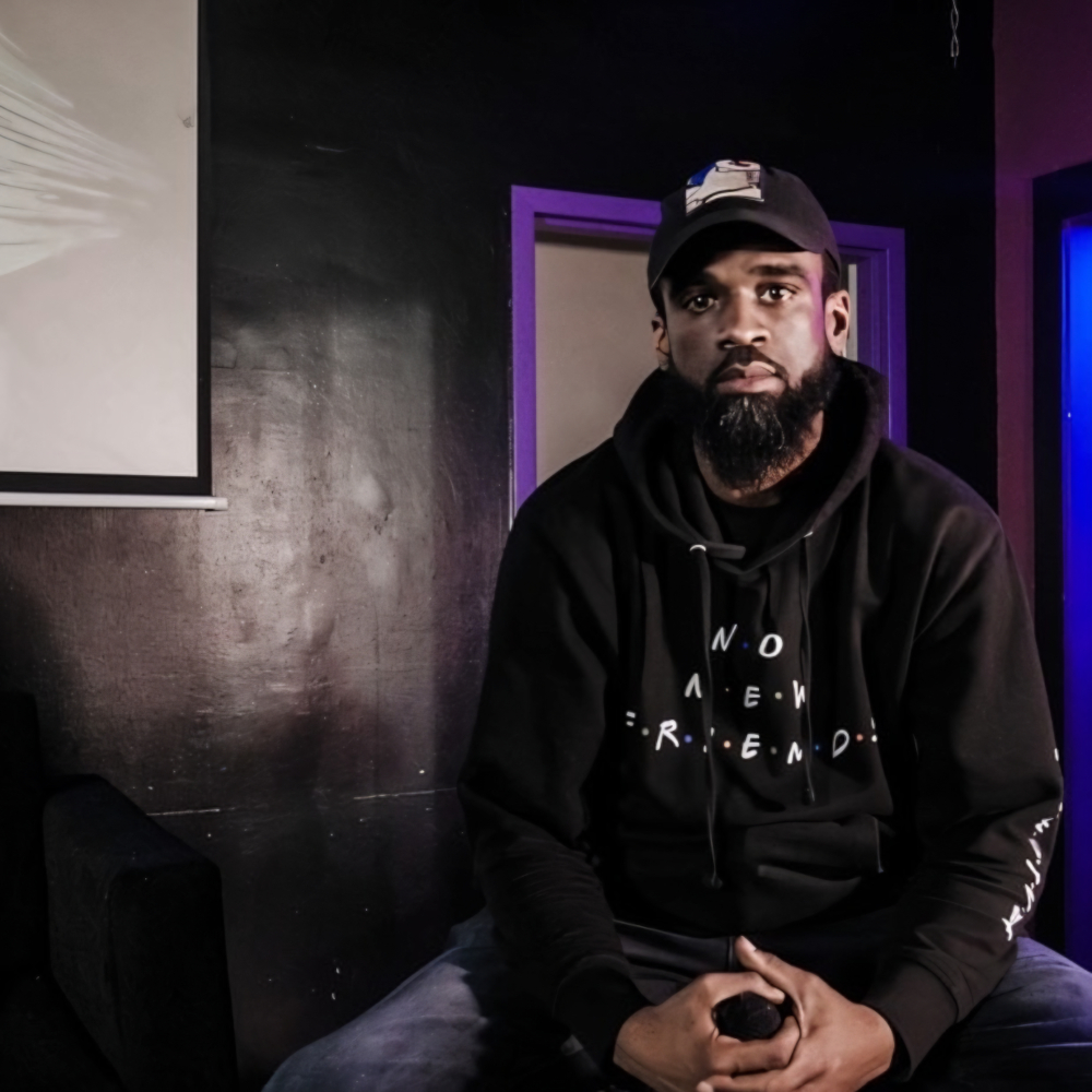 American rapper and producer Classical The Great is sitting with his hands in front of him, wearing a hoodie and baseball cap, and looking directly at the camera. He is not centered in the frame, with a whiteboard and a visible doorway behind him in this color image.