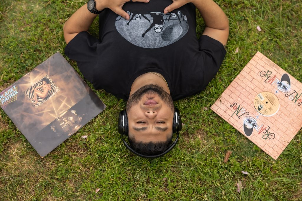 American DJ and radio presenter DJ Nocturne is featured in a vibrant portrait, captured upside down as he lies on the grass with his head facing downward. Two vinyl records are positioned on either side of his head. He wears headphones and a black t-