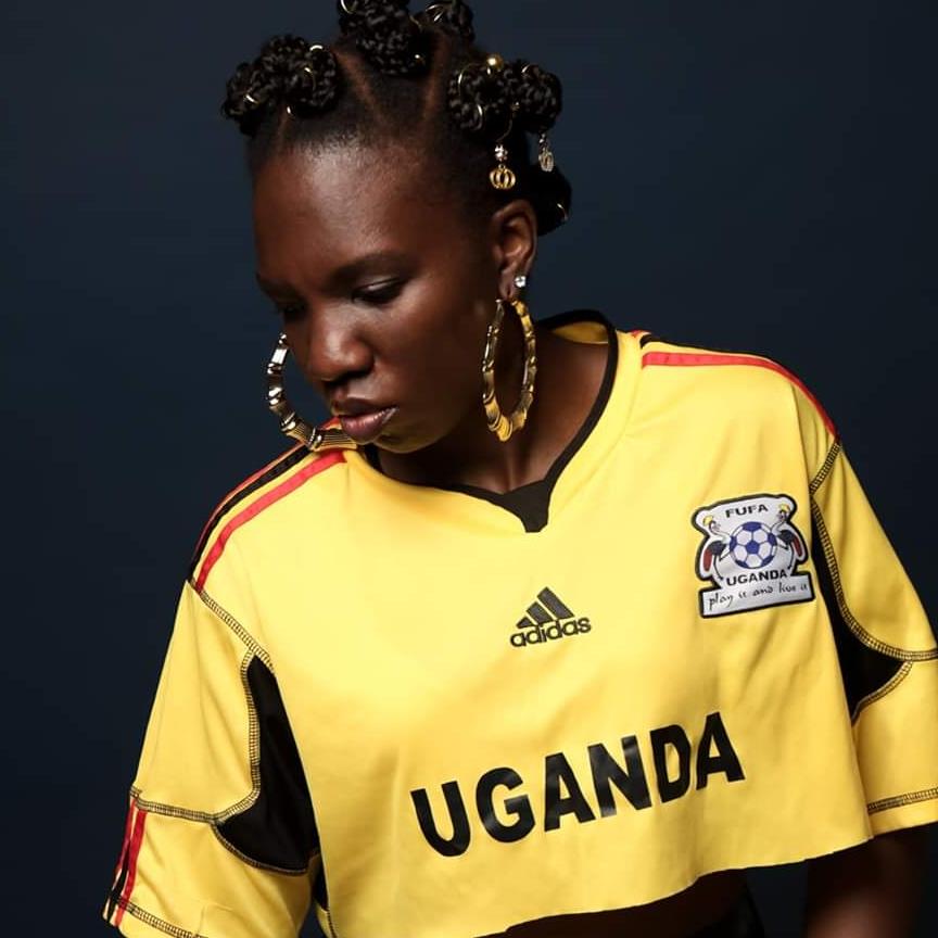 Ugandan-American rapper and activist Emma Lee MC stands looking down to her right, wearing a Ugandan football team shirt, large earrings, and a braid in her hair, captured in a color image.