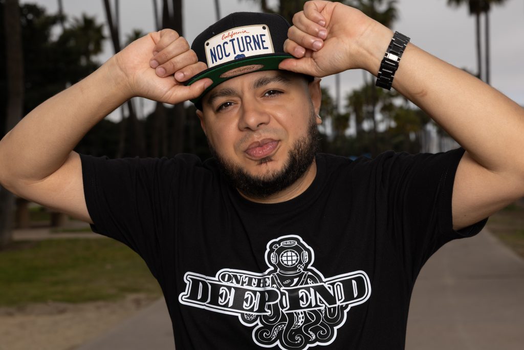 American DJ and radio host DJ Nocturne is captured in a vibrant portrait, looking at the camera with a playful smirk. He sports a DJ Nocturne baseball cap, gripping the brim with both hands. The setting for the photo is Los Angeles, California, USA.