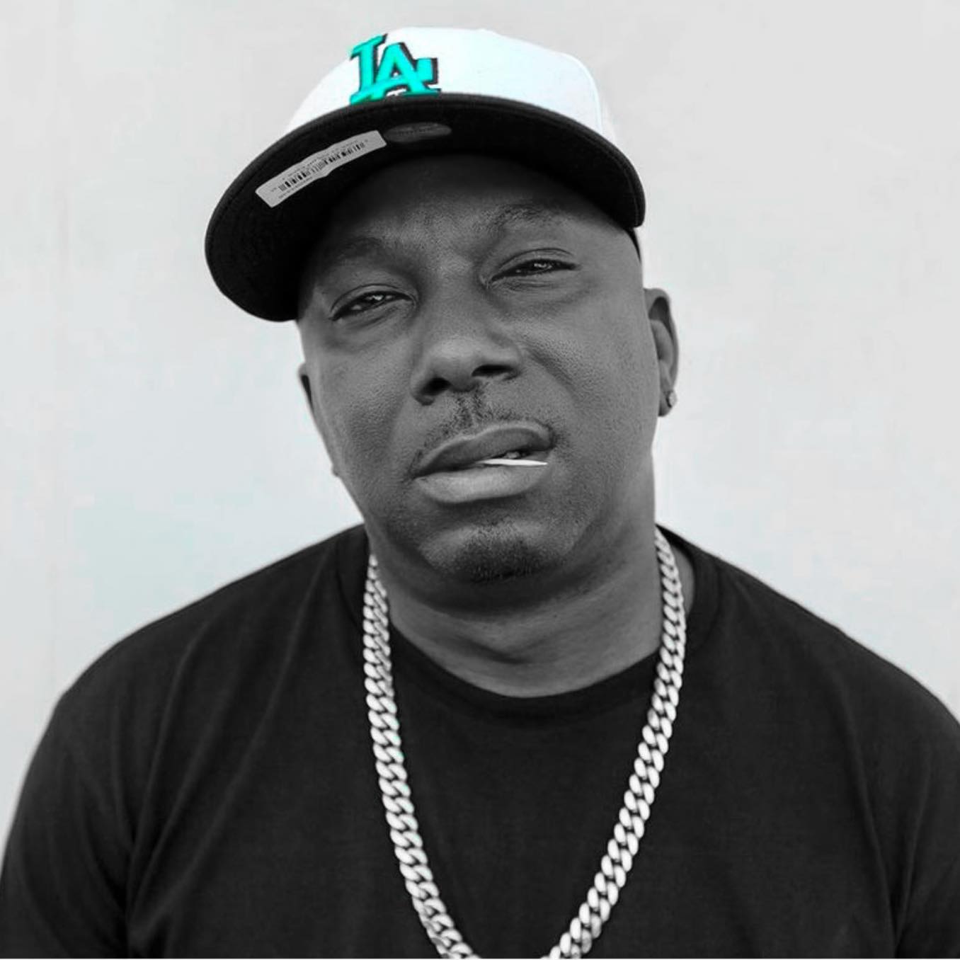 In the image, American rapper Ras Kass sits deep in thought, fixing the camera with a menacing gaze. He’s sporting an LA Dodgers cap with bold blue lettering, a black T-shirt, a gold chain round his neck, and a toothpick between his lips. The shot is vibrant yet tinged with greyscale, with the cap’s lettering standing out in subtle color.
