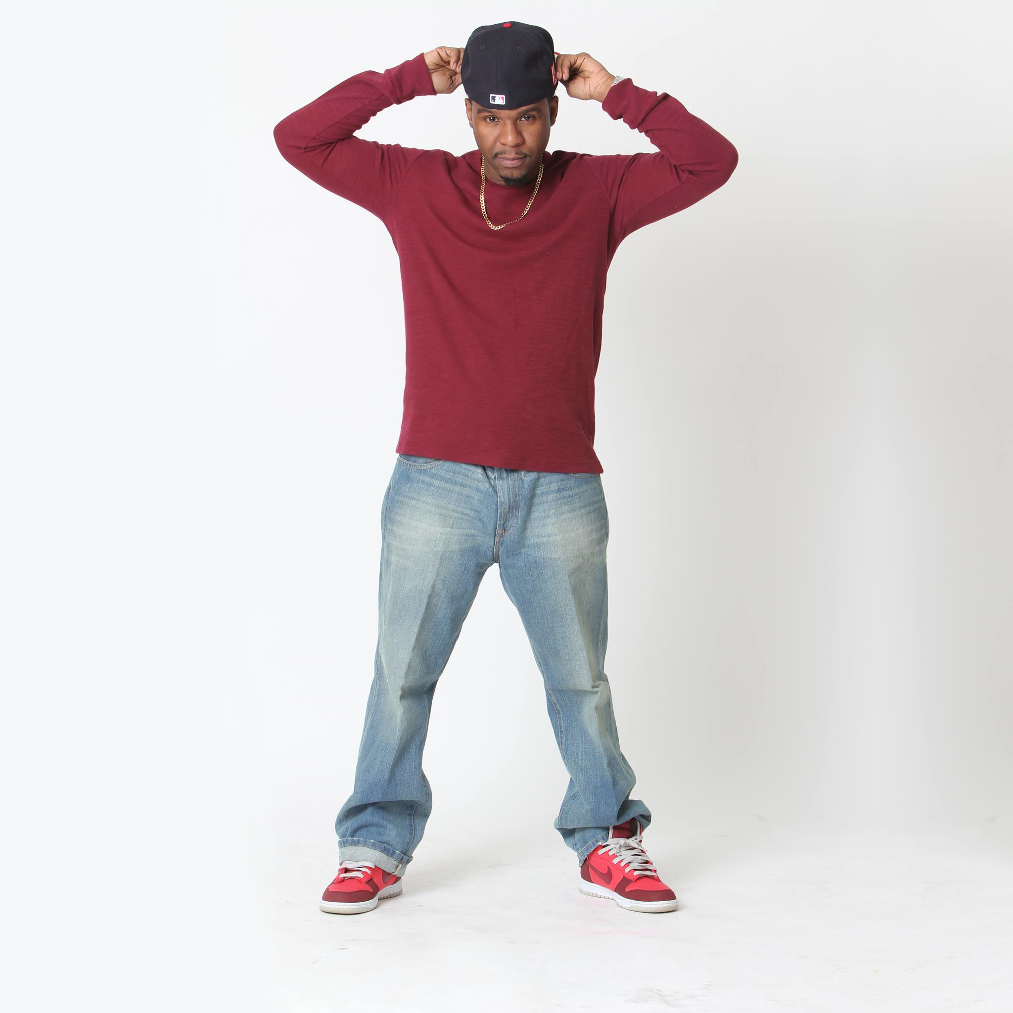 In the image, American rapper Contageous Funk faces the camera wearing a black baseball cap, a burgundy long-sleeved top, light blue denim jeans, and burgundy and pink Nike sneakers. He holds the baseball cap backwards with both hands while standing against a grey and white background. The whole scene is vibrant and full of color.