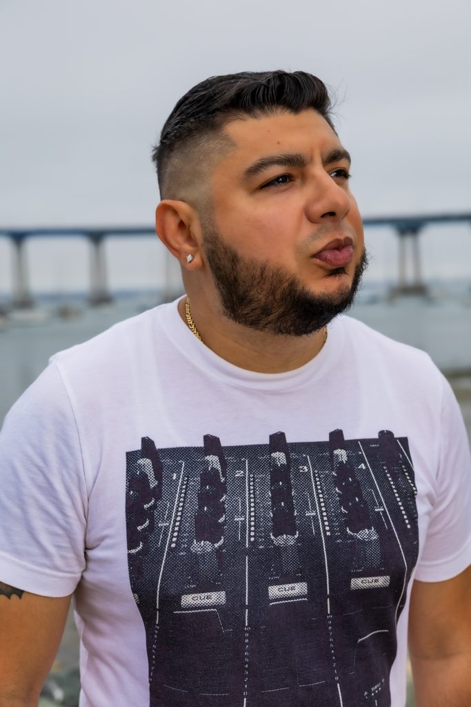 American DJ and radio presenter DJ Nocturne is captured in a vibrant portrait, standing near the harbor and gazing to his left. He’s wearing a t-shirt featuring a Pioneer DJM800 mixer printed on white fabric, with the lively scene set against the bac