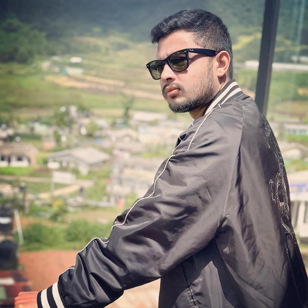 Sri Lankan DJ and producer Maysot stands on a balcony, gazing to his left with his back to the camera, dressed in a black jacket and wayfarer sunglasses, with fields stretching out in the background on a sunny day in this colour image.