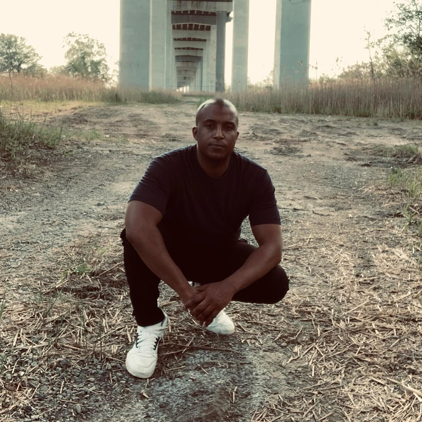 American-born Canadian rapper Dialect Sosa is crouching under a raised road bridge, gazing at the camera in a color image, dressed in white sneakers, black jeans, and a black T-shirt.