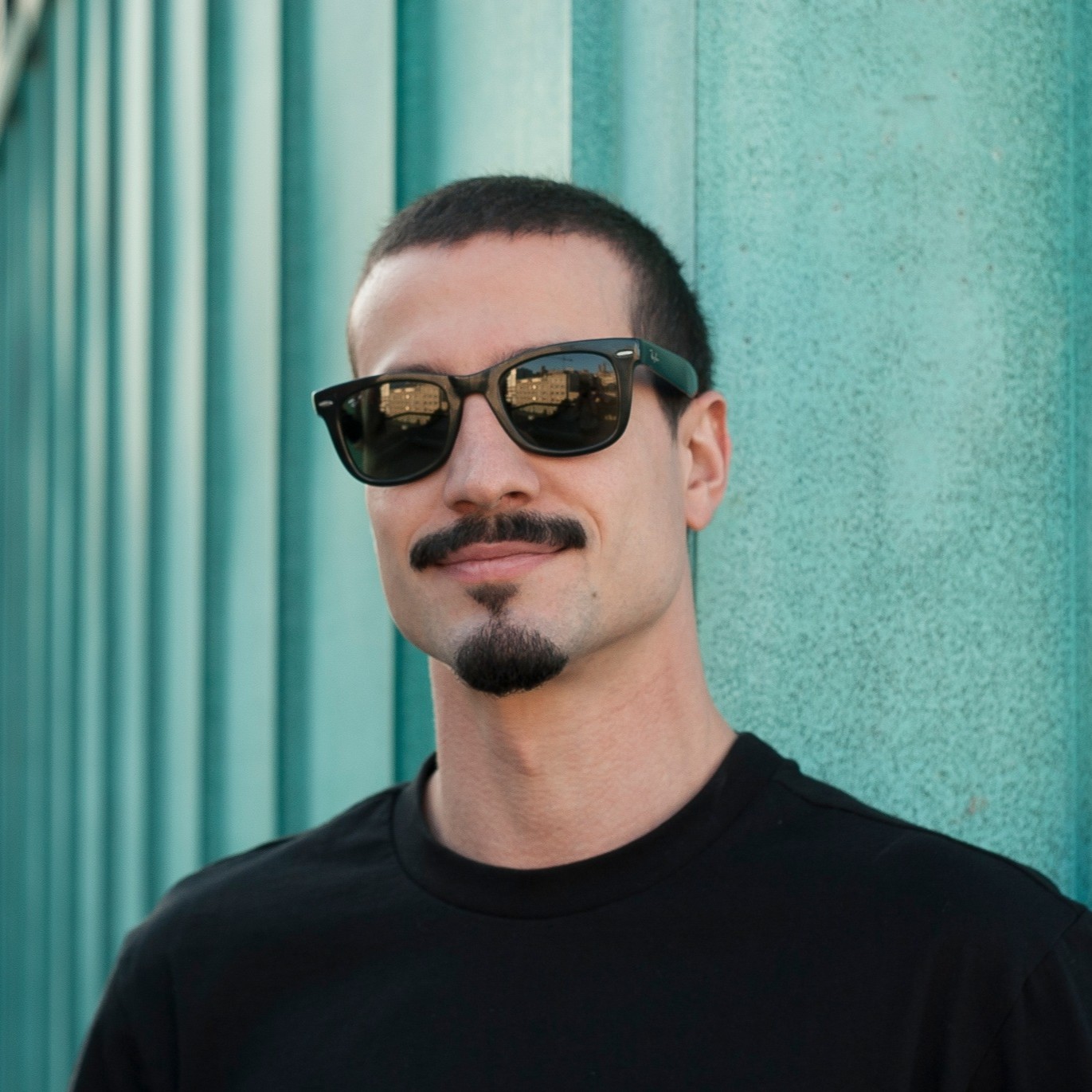 Argentinian DJ and producer Plafnik stands in front of a green container, smirking while wearing sunglasses, a black T-shirt, and sporting a goatee beard and mustache in a color shot.