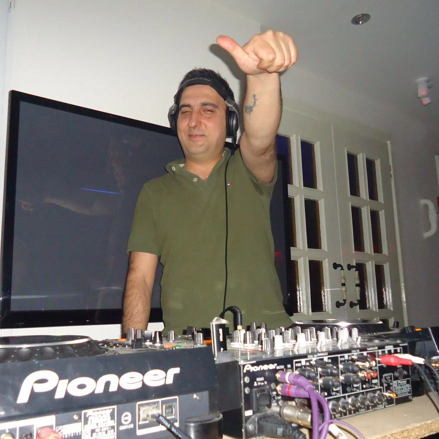 Iranian DJ, producer and label owner Ali Pink performing behind Pioneer DJ equipment, wearing headphones and raising one hand during a club set. Based in Tehran, he is the founder of Techno Tehran Records and supports underground electronic music artists from Iran and the wider Middle East.