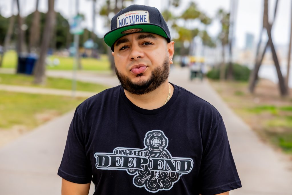 American DJ and radio host DJ Nocturne is pictured in a vibrant landscape shot, gazing at the camera with a playful smirk. He wears a DJ Nocturne baseball cap and an “On the Deep End” T-shirt, with both arms relaxed at his sides. The photo is set in
