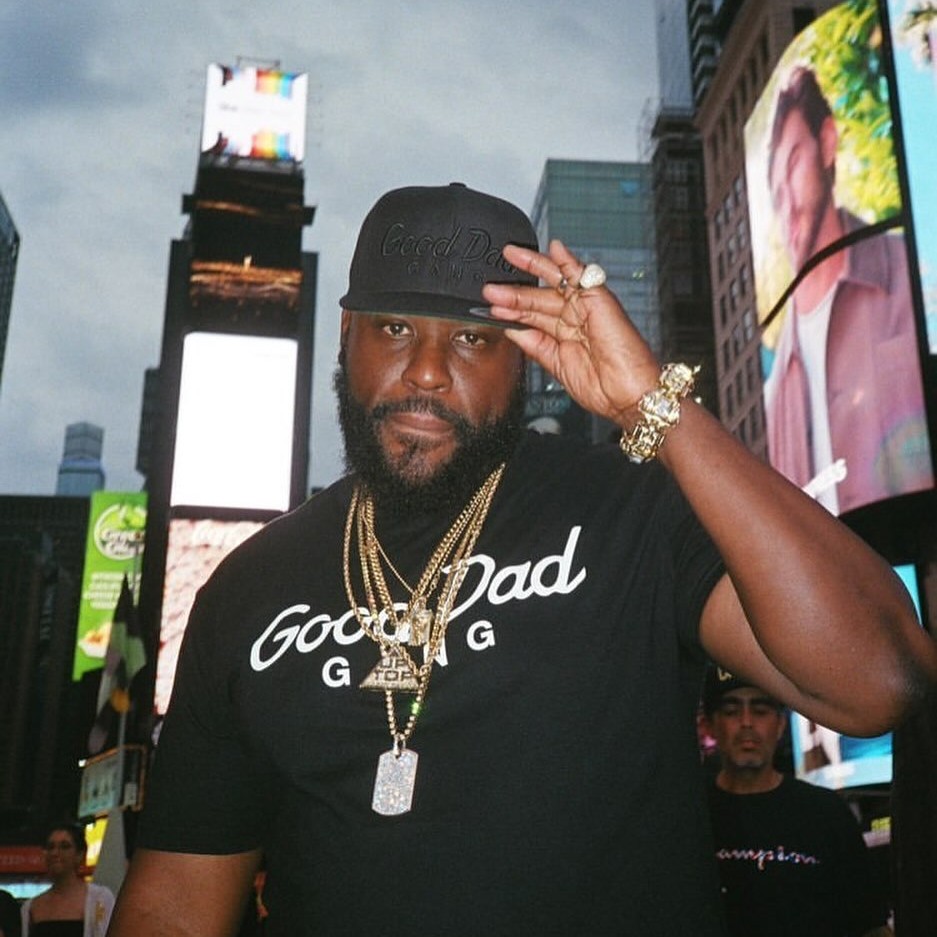In the image, American rapper Wais P faces the camera, wearing a baseball cap with his left hand on the brim and a Good Dad Gang T-shirt for Father’s Day, adorned with multiple gold chains around his neck on a street in New York. The entire scene bursts with vibrant colors and energy.