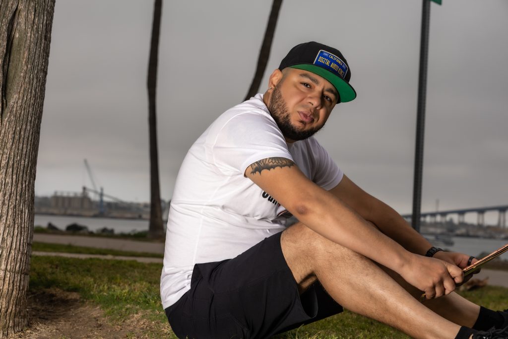 American DJ and radio host DJ Nocturne is captured in a lively outdoor scene, sitting under a tree with his body turned to the left, glancing right at the camera with a playful smirk. Sporting a DJ baseball cap and a “Cali's HouseNation Radio” T-shir